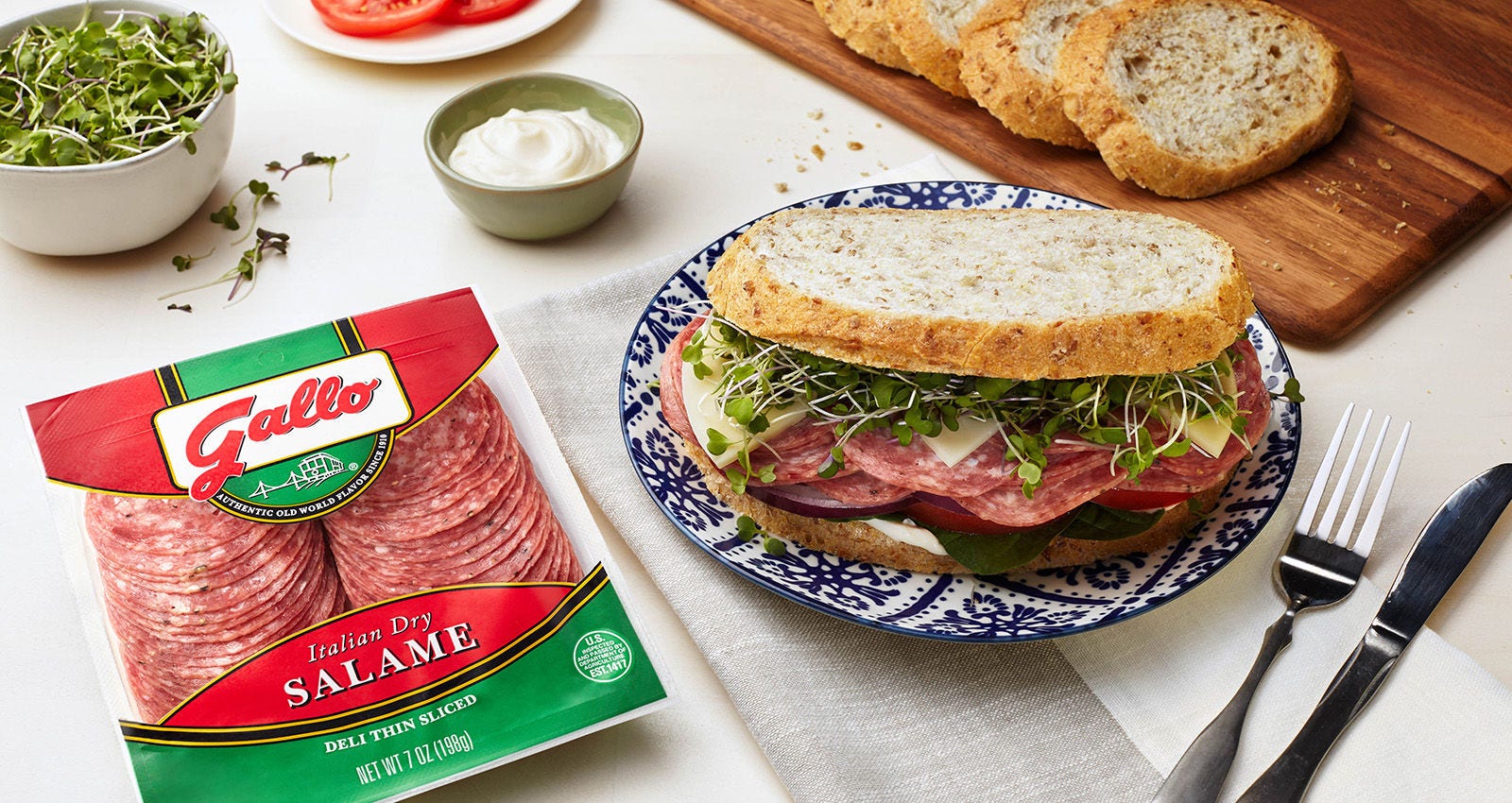 Gallo Italian Dry Salame package next to a stacked salame sandwich with provolone, greens, and crusty bread on a bright tabletop scene